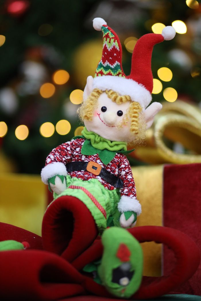 services-01 Charming elf doll seated among Christmas decorations with glowing lights behind.